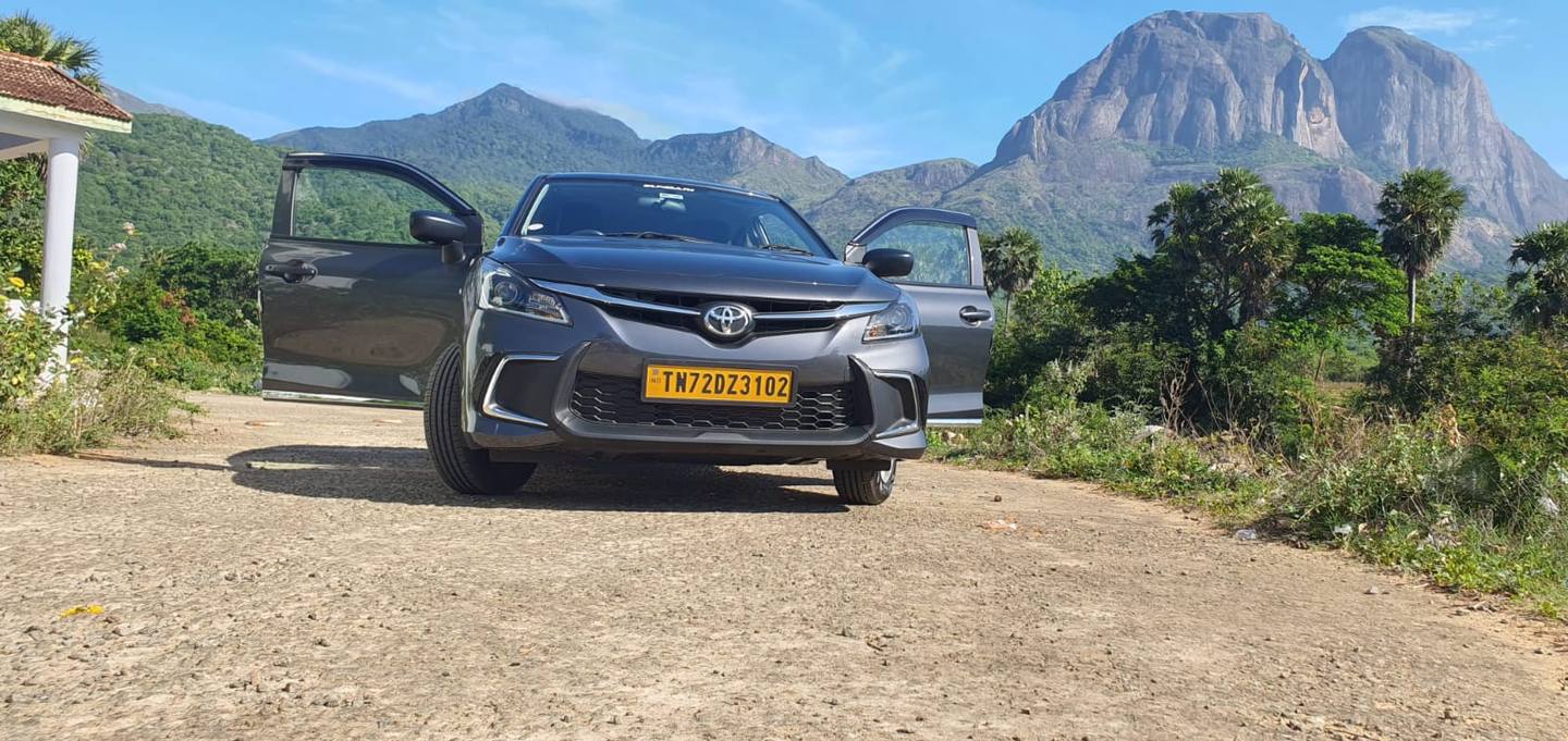 Our grey Toyota Glanza with both front doors open on a gravel road below green hills and a rocky peak