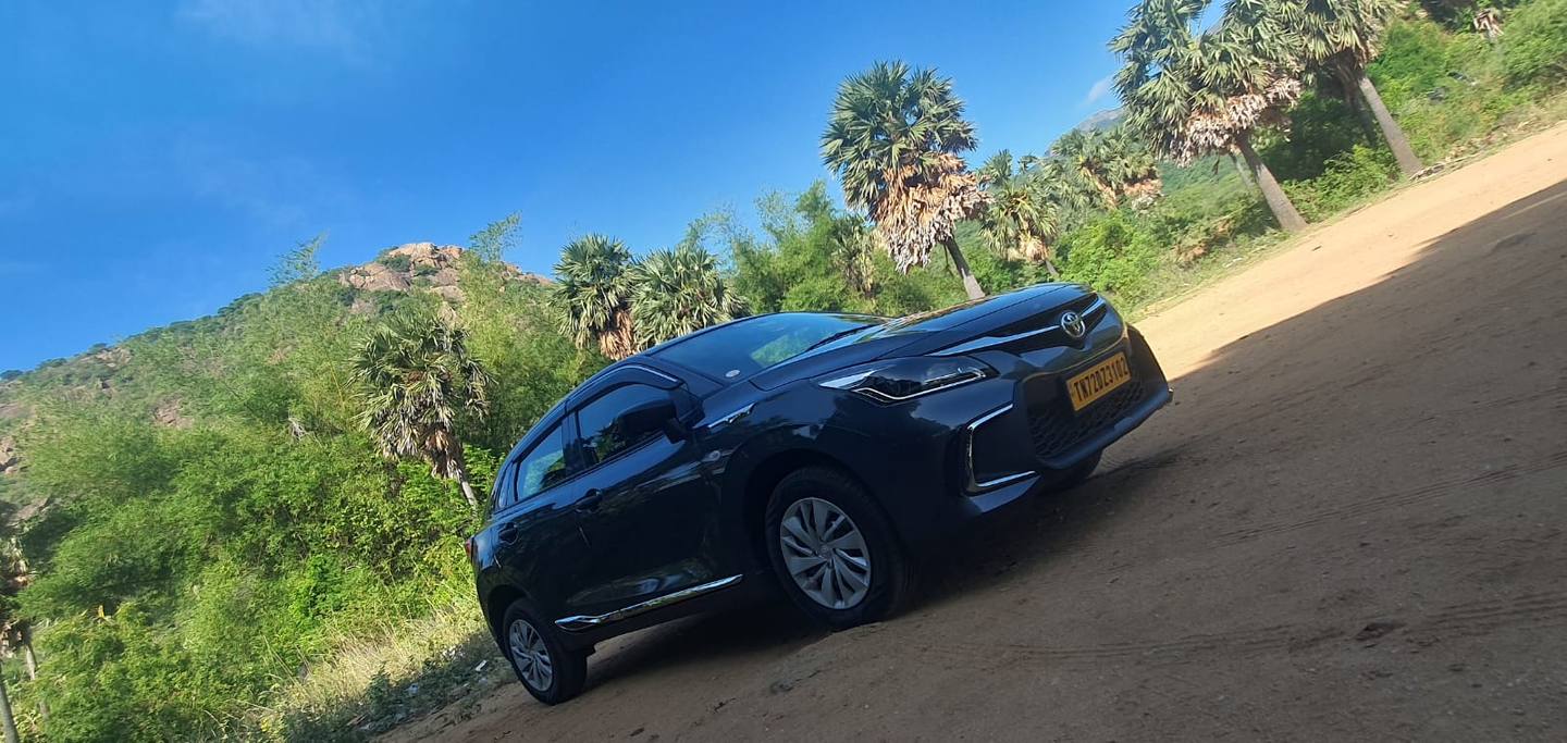 Our Toyota Glanza on a dirt road, with palmyra palms and a green hill behind