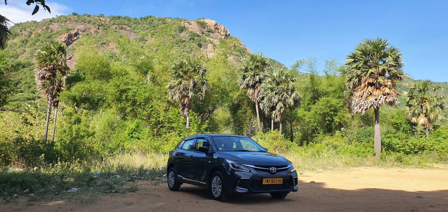 Our grey Toyota Glanza parked under palm trees with a green hill behind